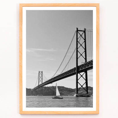 Lisbon Bridge Black and White Poster by Raphael Alves featuring a framed photograph of a suspension bridge and sailboat in the water, framed and displayed in modern living room decor