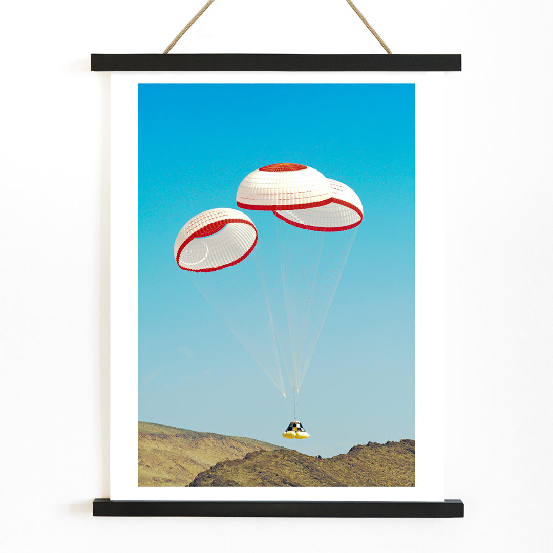 CST-100 crew capsule landing with three parachutes over desert landscape by NASA and The Boeing Company poster artwork, with wooden magnetic frame on white background